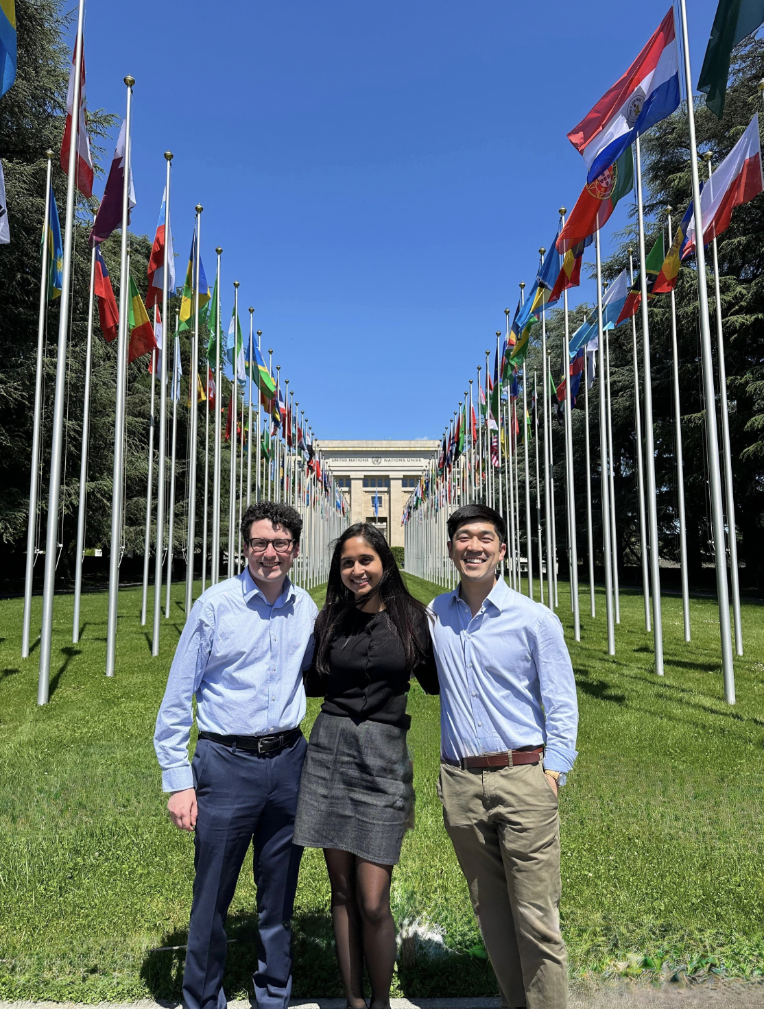 Three people stand together in a grassy area lined with flags from around the world.